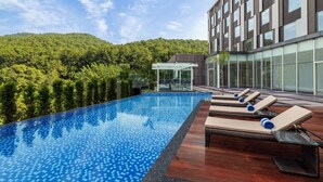 Una piscina infinita en la azotea con sillas de descanso, rodeada por un edificio moderno y una ladera boscosa.