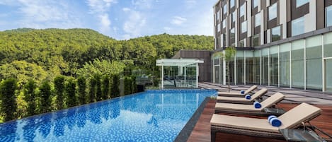 A rooftop infinity pool with lounge chairs, surrounded by a modern building and a forested hillside.