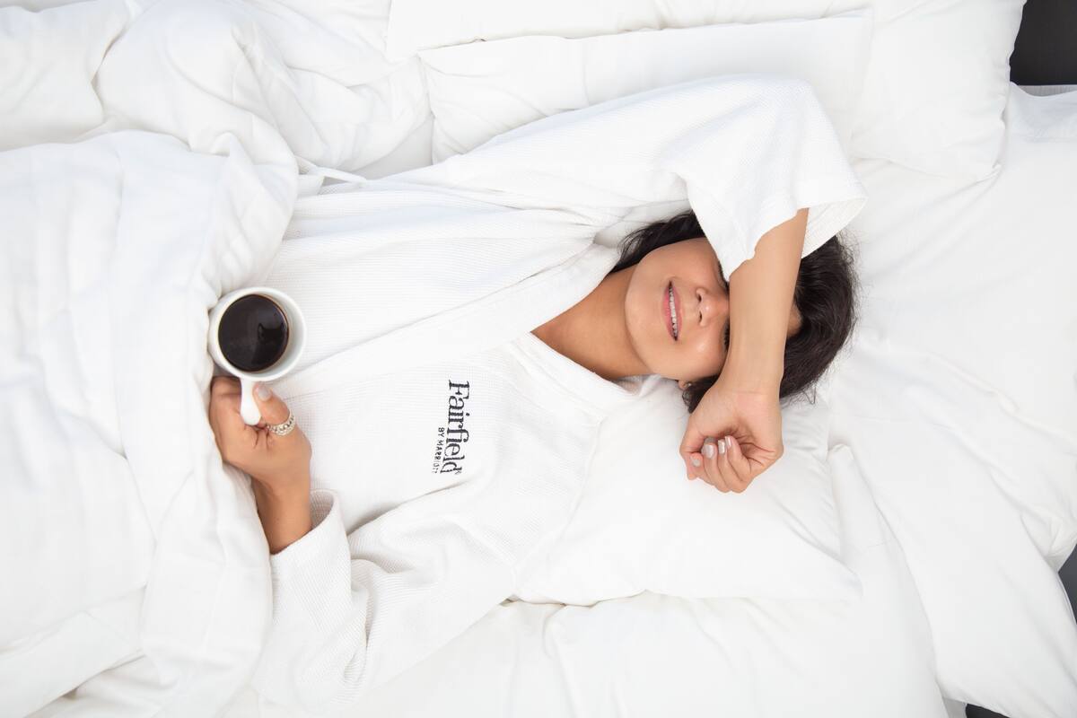 A person lying in bed with a cup of coffee, wearing a white robe.