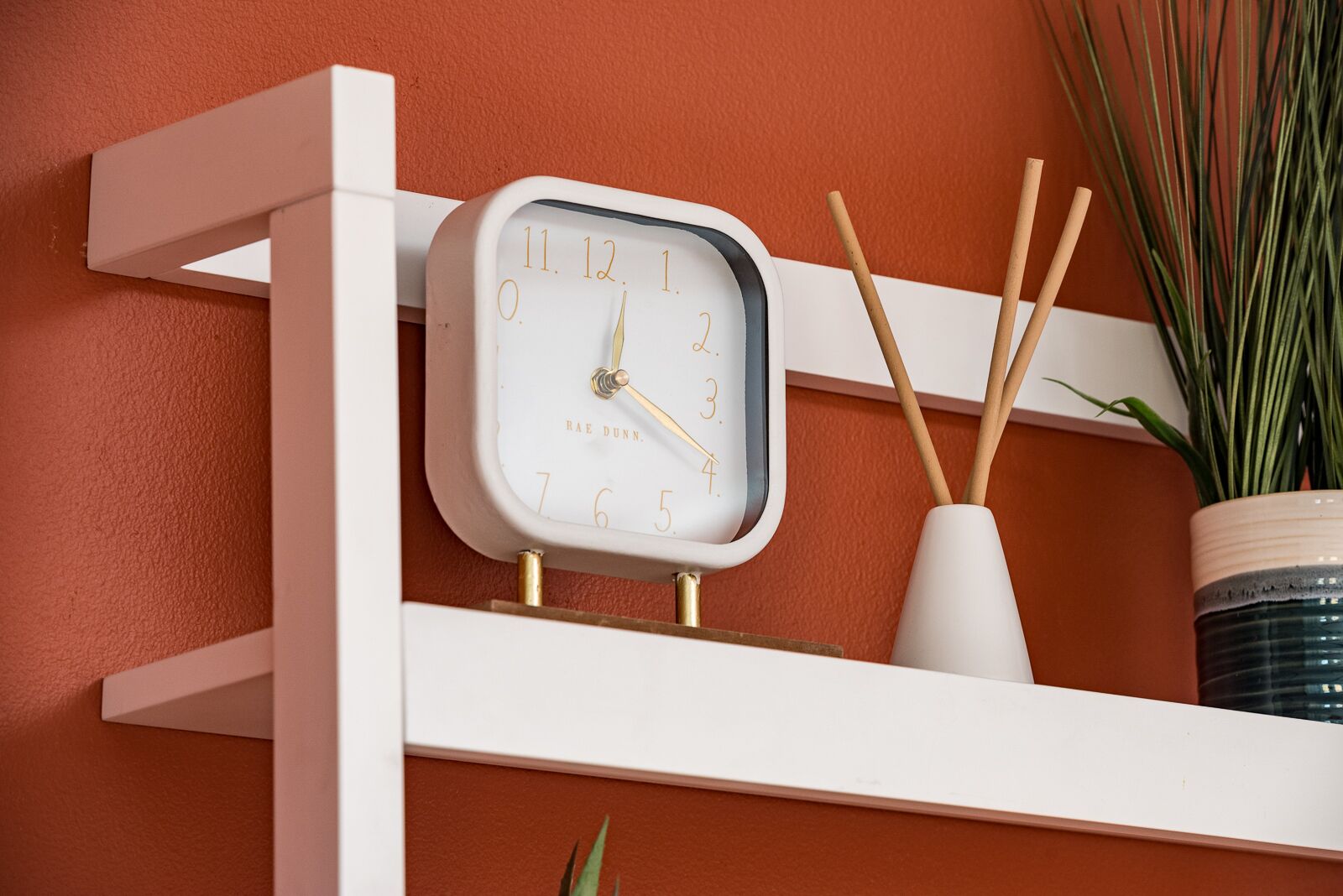 A white clock with gold numbers and hands, mounted on a white shelf against a textured orange wall.