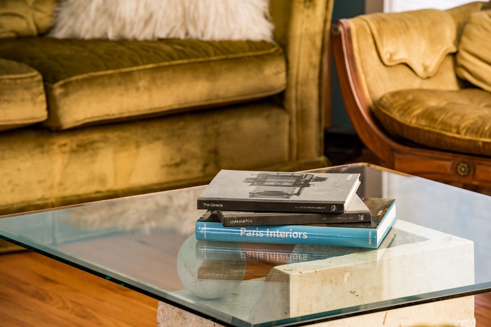 Een stapel boeken op een glazen tafel, met een bank en fauteuil op de achtergrond.