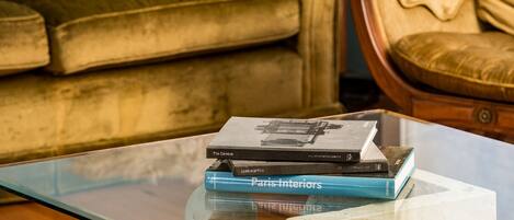 Een stapel boeken op een glazen tafel, met een bank en fauteuil op de achtergrond.
