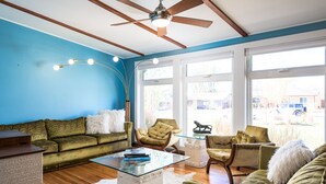Ein Wohnzimmer mit blauer Wand, Deckenventilator, Sofa, Glastisch und Blick ins Freie.