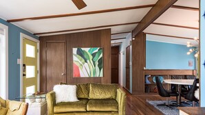 Ein Wohnzimmer mit einem grünen Sofa, einem weißen, flauschigen Kissen, einem Glastisch und einem Essbereich mit weißem Tisch und schwarzen Stühlen.
