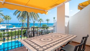 Un balcón con una mesa y sillas de cerámica, con vistas a una piscina y palmeras, y una vista al mar.