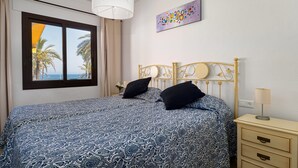 Un dormitorio con cama, mesita de noche, lámpara, un cuadro y ventana con vistas a palmeras y al mar.