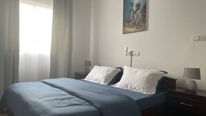 Un letto rifatto con lenzuola blu, un comodino con una lampada e un quadro appeso al muro.