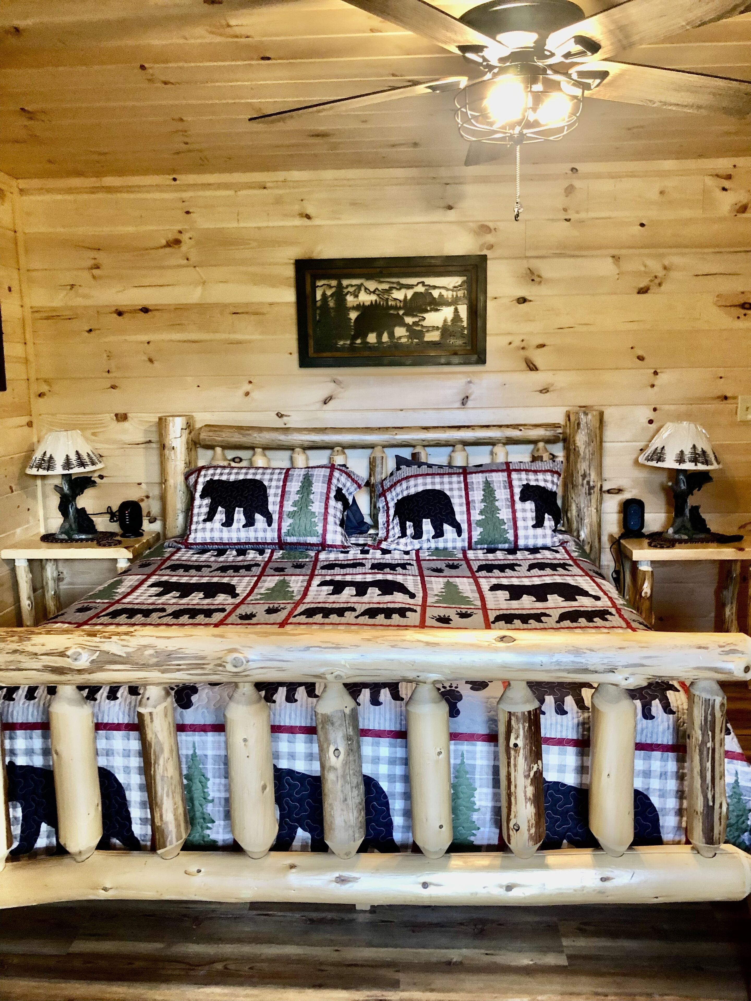 Un dormitorio de cabaña de madera con una cama, un edredón con motivos de oso, dos lámparas de noche y un ventilador de techo.