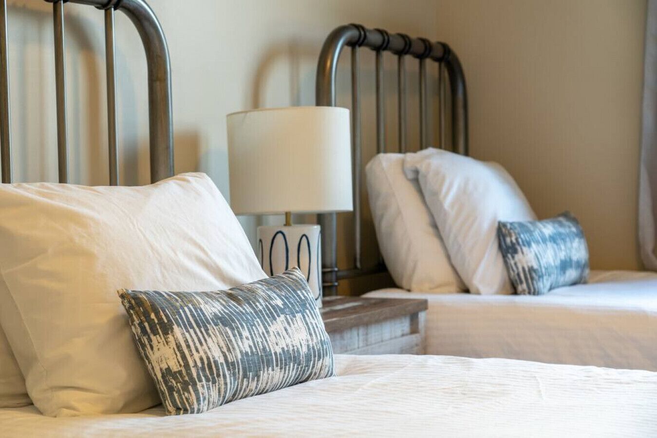 A hotel room with two beds, each with white bedding and patterned pillows. There is a bedside table with a lamp between the beds.