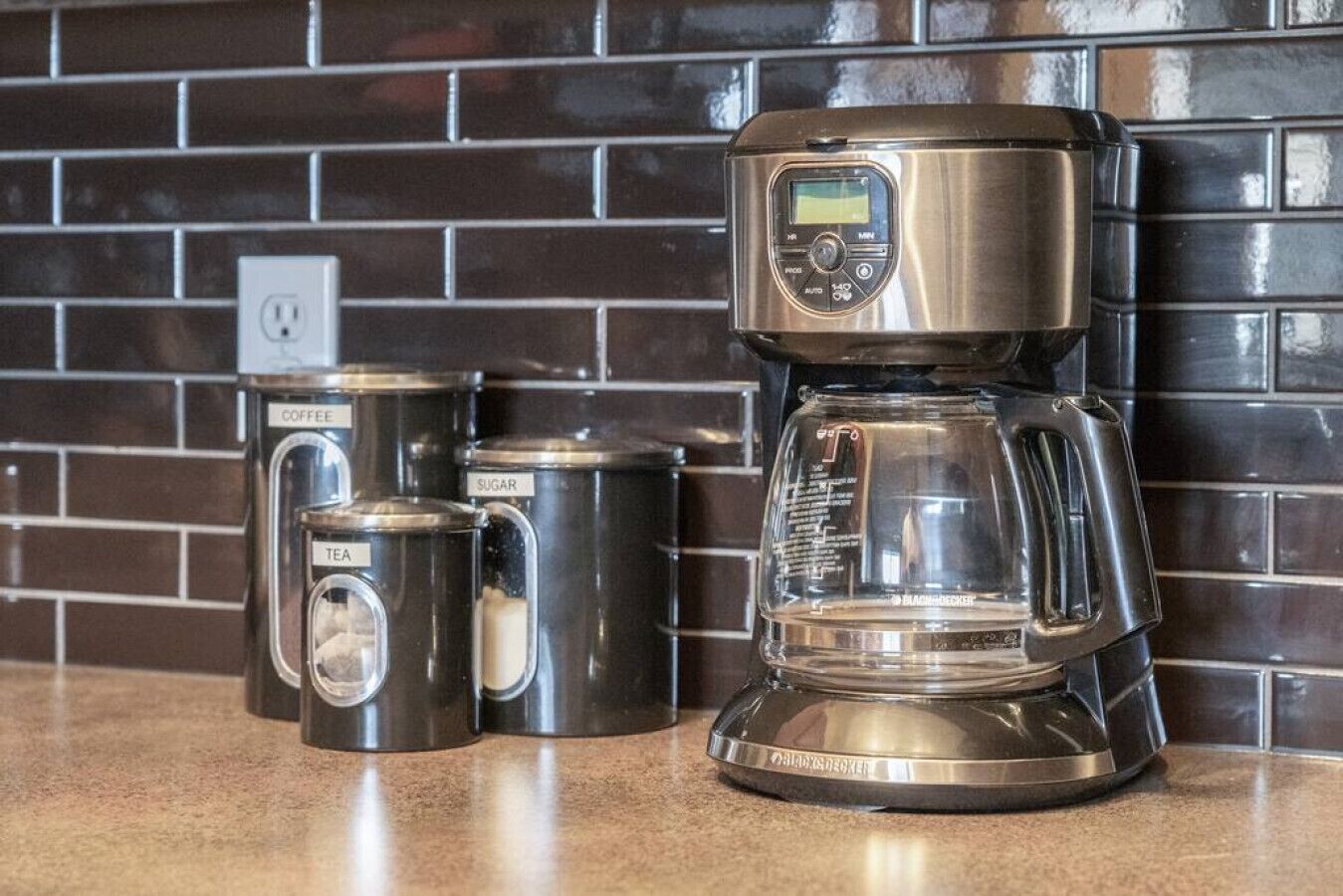 A coffee maker with a digital display, a tea container, and two sugar containers on a kitchen counter.