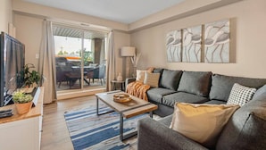 A modern living room with a grey sofa, a coffee table, and a TV. There is a sliding glass door leading to an outdoor area with a table and chairs. - Modern Boho | Mountain Views | Pool | Hot Tub (Invermere)