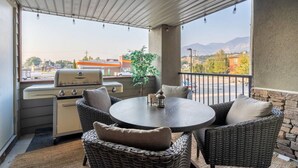 A covered patio with a grill, wicker furniture, and a view of the city and mountains. - Modern Boho | Mountain Views | Pool | Hot Tub (Invermere)