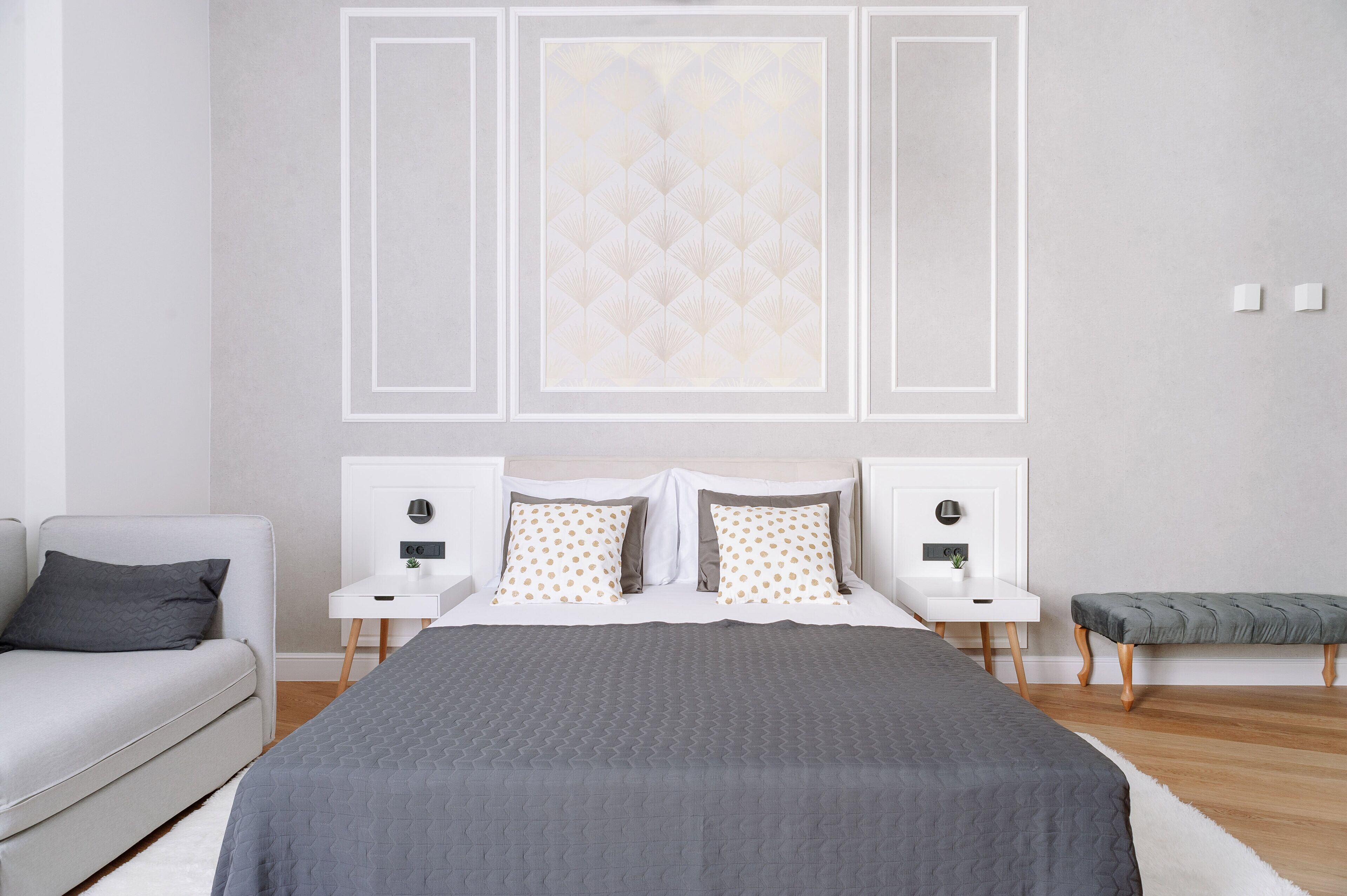 Une chambre moderne avec un grand lit, deux tables de chevet, une commode, un banc gris et un mur blanc orné de panneaux décoratifs.