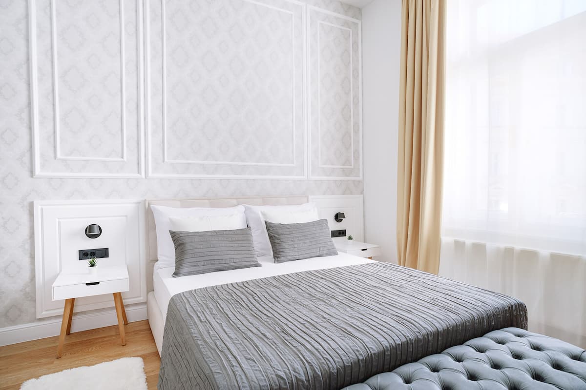 A bedroom with a bed, two bedside tables, a nightstand, a window with curtains, and a white wall with a subtle pattern.