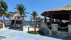 A beachside restaurant with a thatched roof, a fenced outdoor seating area, and palm trees in the background.