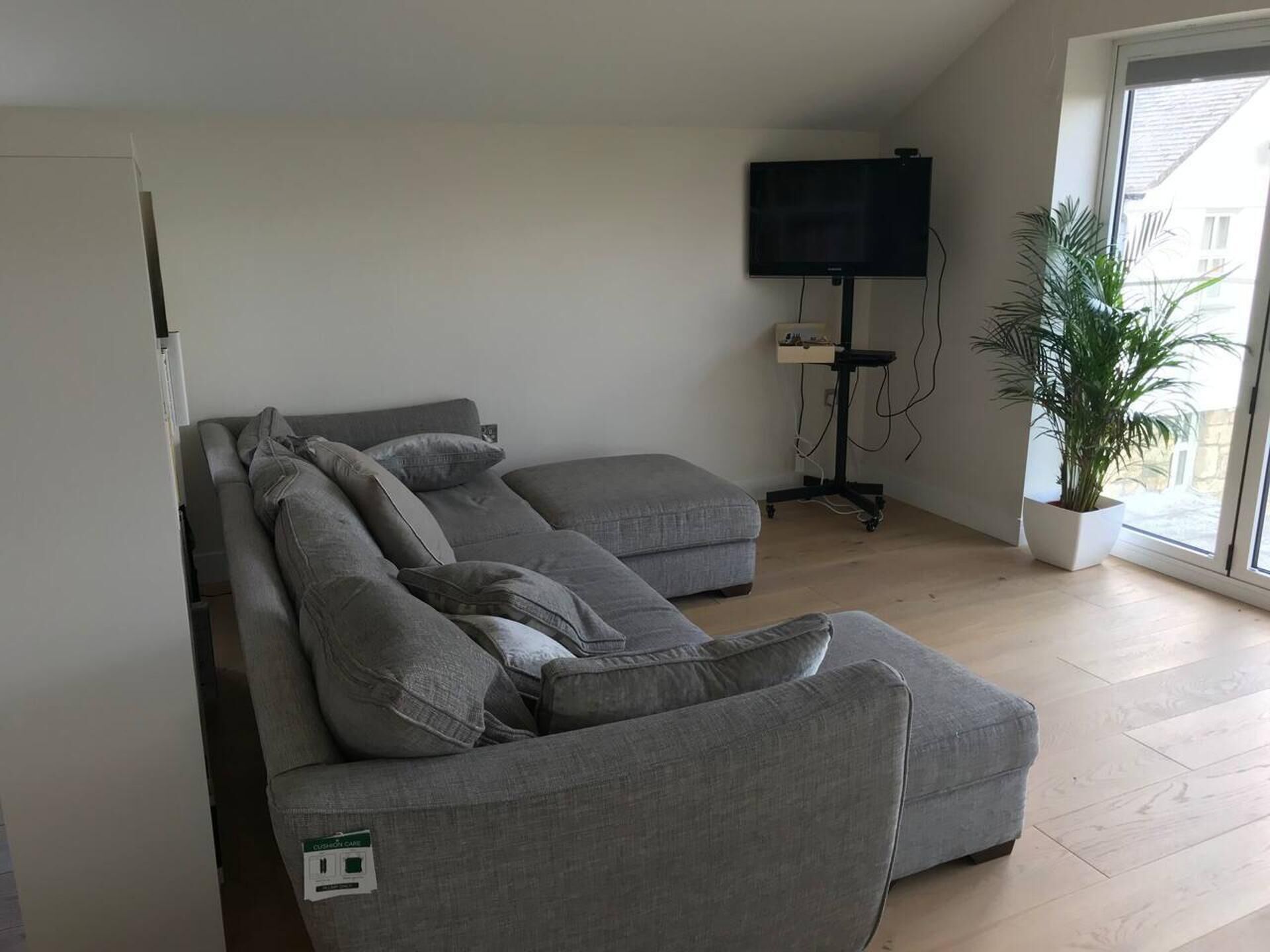 A modern living room with a grey sectional sofa, a flat-screen TV mounted on the wall, a small table with a plant, and a large window providing natural light.