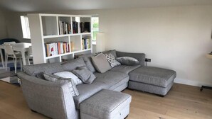 A grey sectional sofa with a matching ottoman, a white bookshelf, and a dining area with a white table and chairs.
