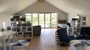 A spacious room with a sloped ceiling, a large bookshelf, a desk, a bed, and a view of greenery through large windows.