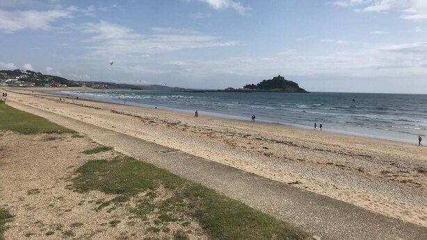 A beach with a grassy path, a sandy shore, and a distant hill with a structure on top.