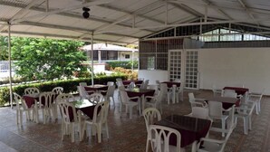 Uma área de refeições coberta ao ar livre, com cadeiras e mesas de plástico branco, dispostas em fileiras.