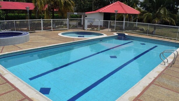 Piscina retangular com marcações azuis para pistas de natação, cercada por uma cerca branca e um edifício com telhado vermelho.