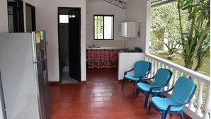 Cozinha compacta com geladeira, pia e janela. Há cadeiras azuis numa varanda com vista para o verde.