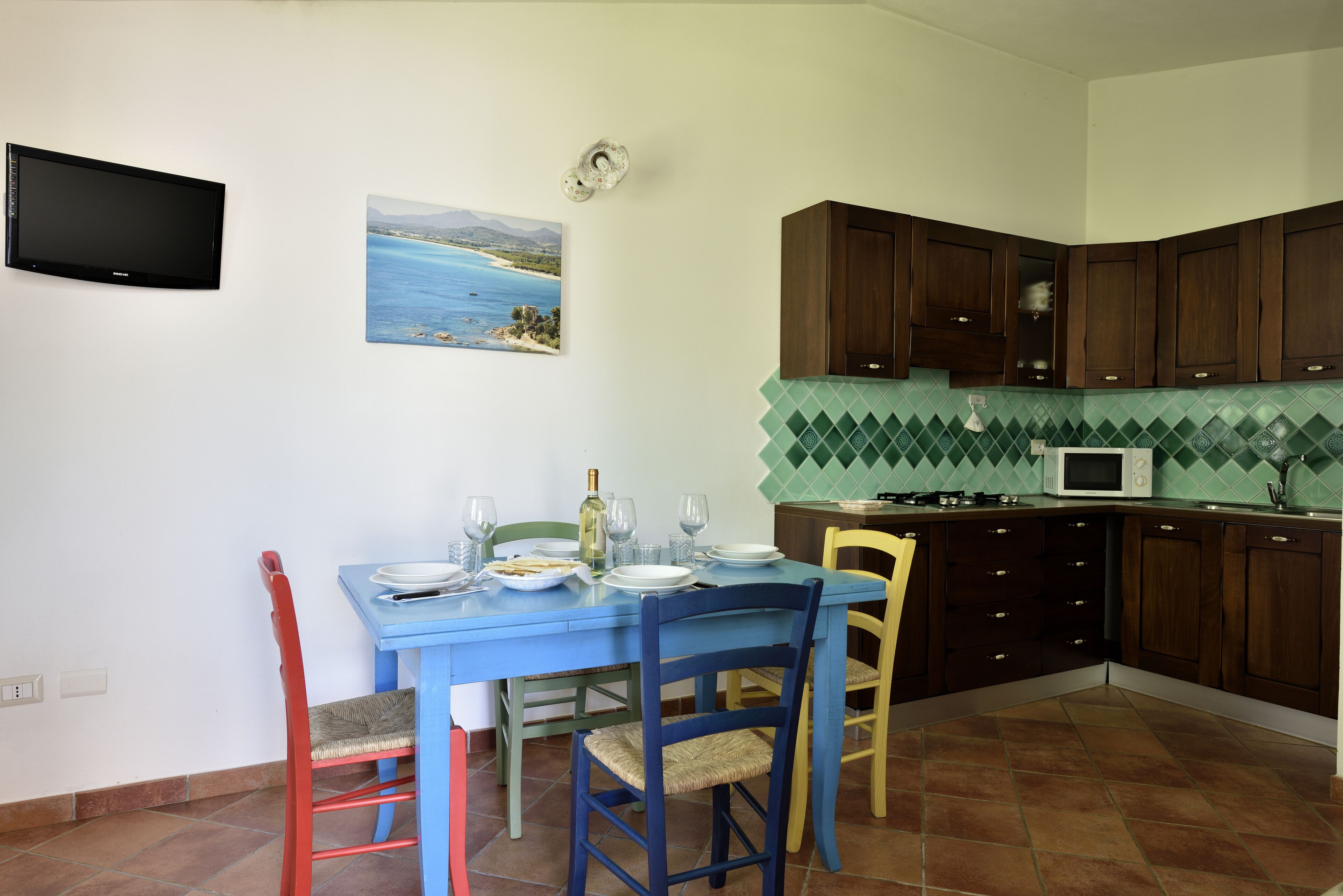 Un coin repas avec une table dressée pour deux personnes, un téléviseur fixé au mur et une cuisine dotée de meubles en bois et d’un plan de travail carrelé.