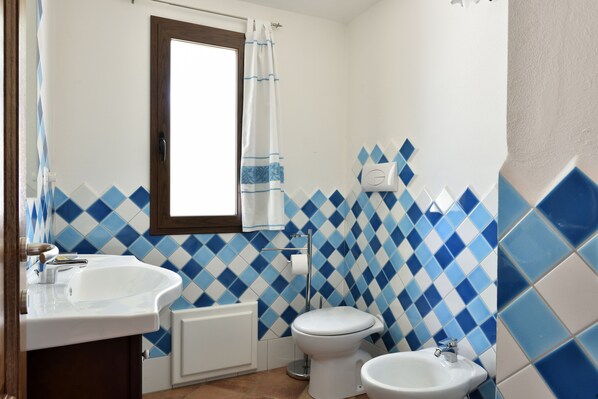 A bathroom with a white sink, toilet, and bidet, blue and white patterned tiles, and a window with a curtain.