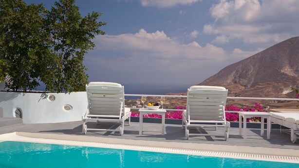 Un espace aménagé au bord de la piscine, avec deux chaises longues blanches, une table sur laquelle se trouvent un saladier rempli de fruits et une bouteille, et une vue sur la mer et les montagnes.