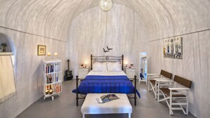 Une pièce avec un plafond incurvé, un lit recouvert d’une couverture bleue, une bibliothèque blanche, un banc et une guitare.
