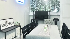 A modern dining area with a long table, black chairs, a grey sofa, a TV mounted on the wall, and a 'welcome' sign on a shelf. - Brilliant Bungalow (Dayton)