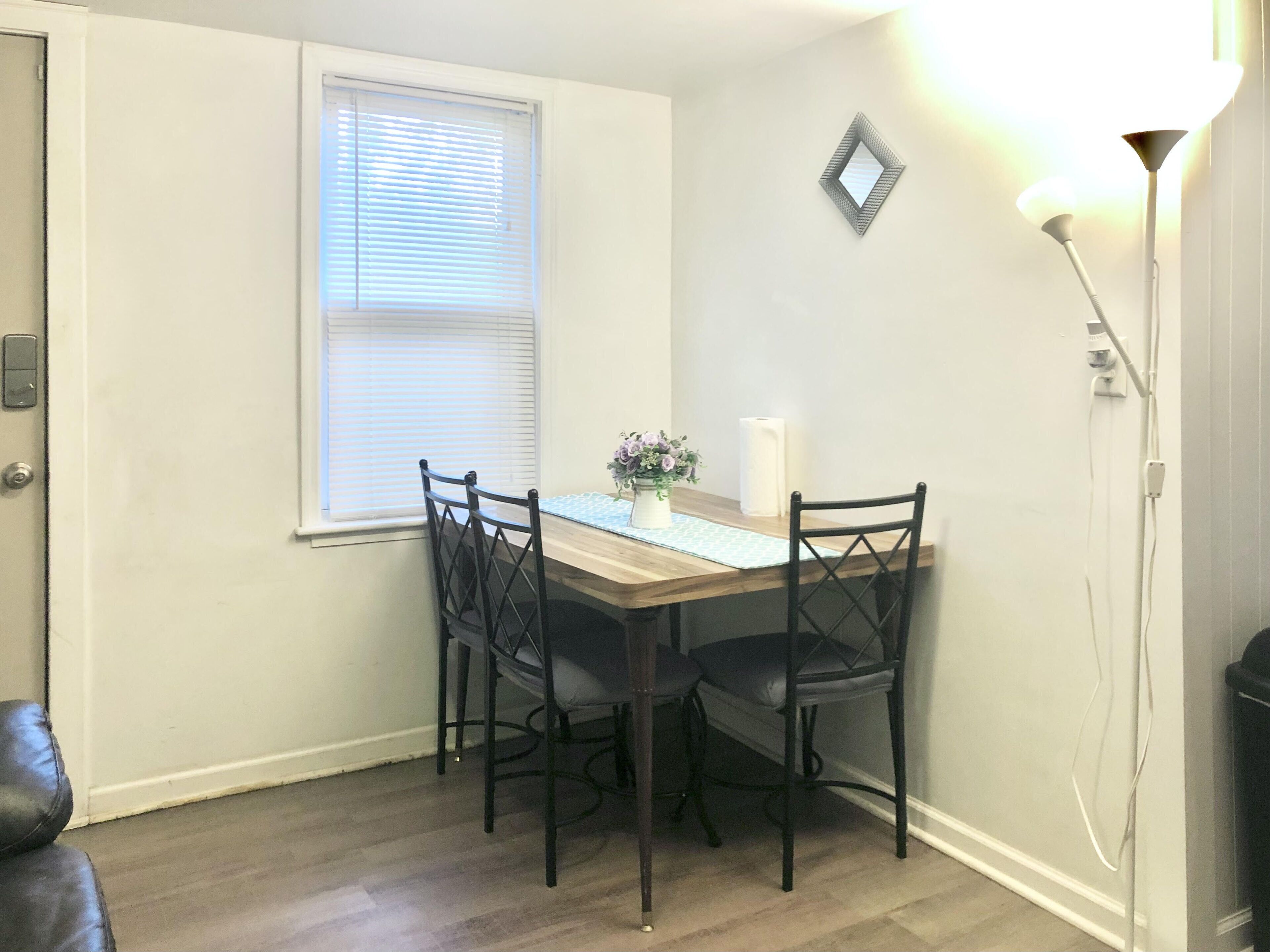 Un coin repas avec une table en bois, quatre chaises, un vase de fleurs, une fenêtre avec des stores et un luminaire fixé au mur.