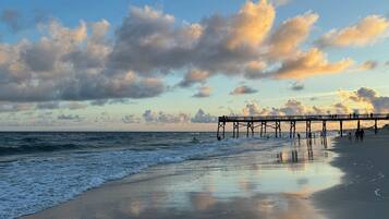 A beach with a pier extending into the ocean, waves gently lapping at the shore, and a partly cloudy sky.