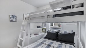 A bunk bed room with a ladder, a shelf with books and a small plant, and a window with blinds.