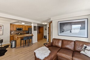 Una cocina moderna con armarios de madera, un refrigerador de acero inoxidable y una zona de bar. Hay un sofá de cuero marrón con una manta de piel y una almohada. Un cuadro grande enmarcado cuelga en la pared.