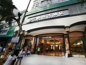 Entrada de hotel con fachada de vidrio, zona de recepción visible y un letrero con caracteres chinos.