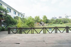 Una terraza de madera con barandilla, rodeada de vegetación y edificios al fondo.