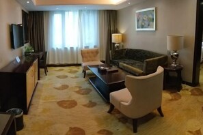 A hotel room with a sofa, two armchairs, a coffee table, a TV mounted on the wall, a desk with a lamp, and a patterned carpet. - Lianhua Hotel (Xiangcheng)