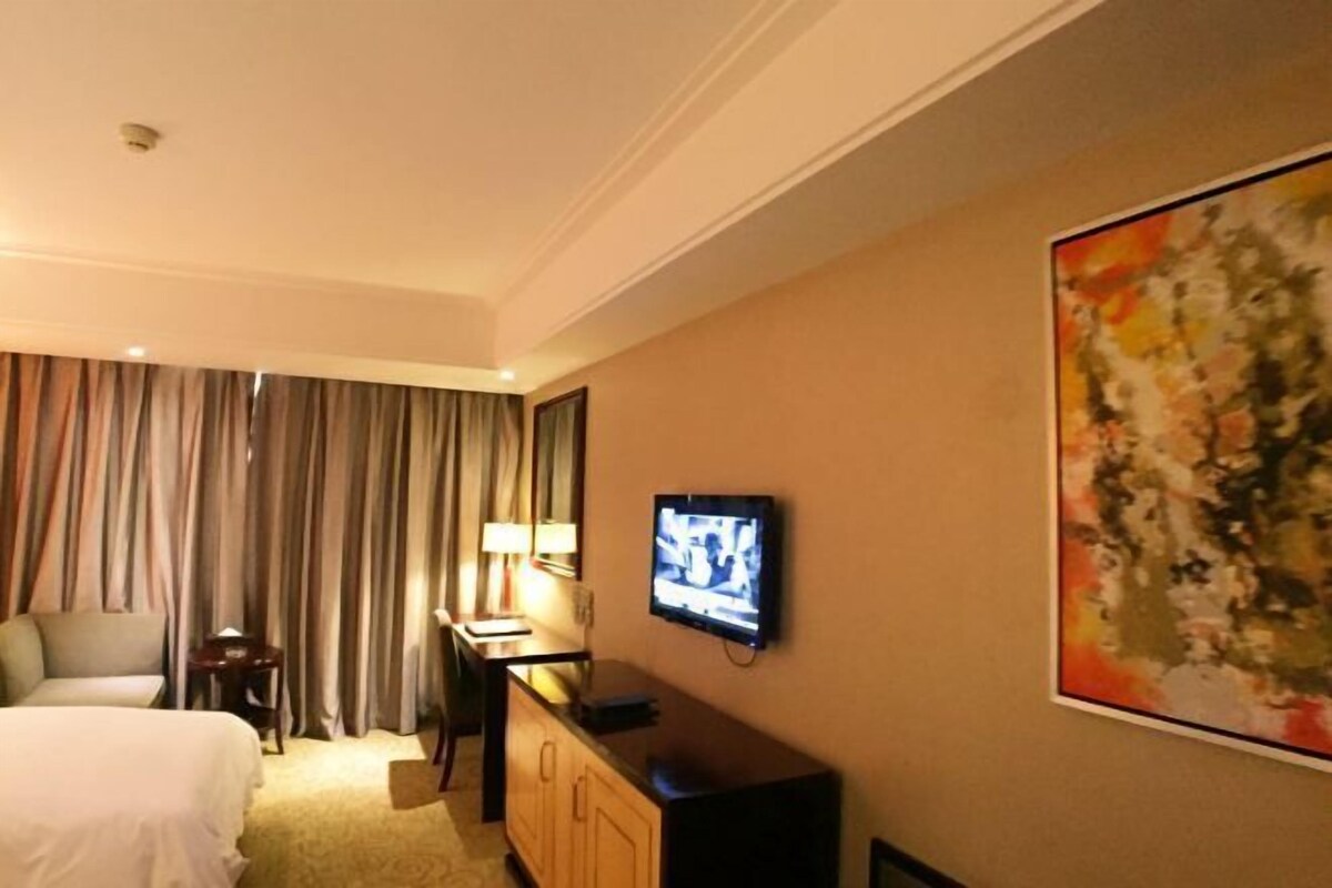 A hotel room with a bed, a desk, a television, and a painting on the wall.