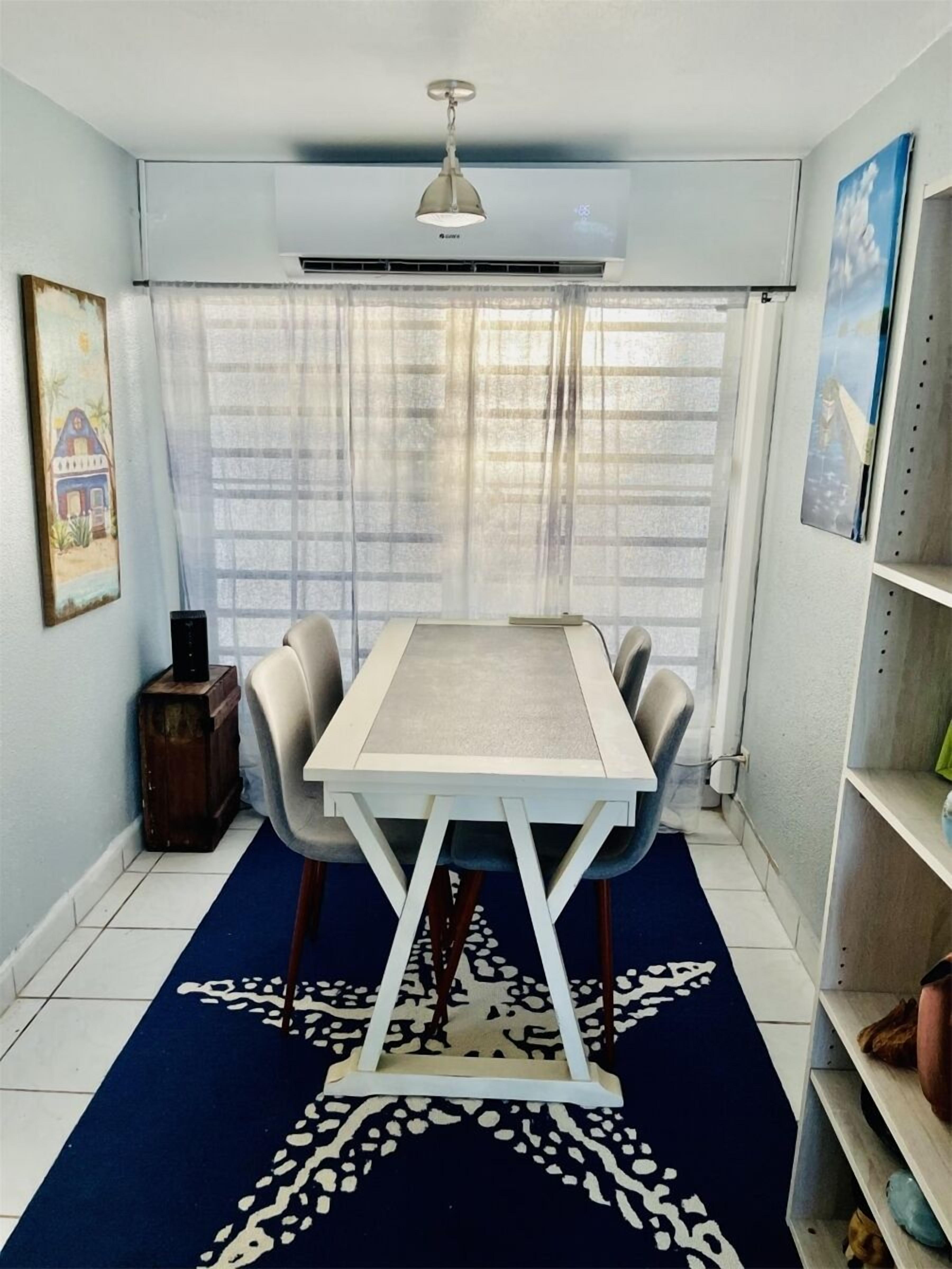 A dining area with a table and chairs, a patterned rug, a ceiling fan, and a window with curtains.