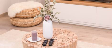Une table ronde en bois, ornée d’un vase de fleurs et de bougies, avec deux télécommandes et un coussin blanc moelleux posé sur un tapis de couleur claire.