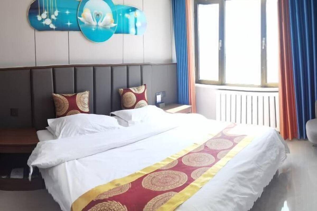 A hotel room with a large bed, a headboard, a bedside table, a nightstand, a window with curtains, and a wall-mounted artwork.