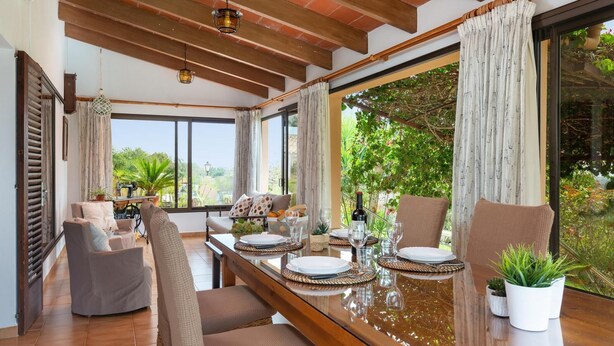 A dining area with a long wooden table set for two, large windows with curtains, and a view of greenery outside.
