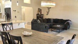A modern living room with a dining area, featuring a wooden table, dark upholstered sofas, and a chandelier.