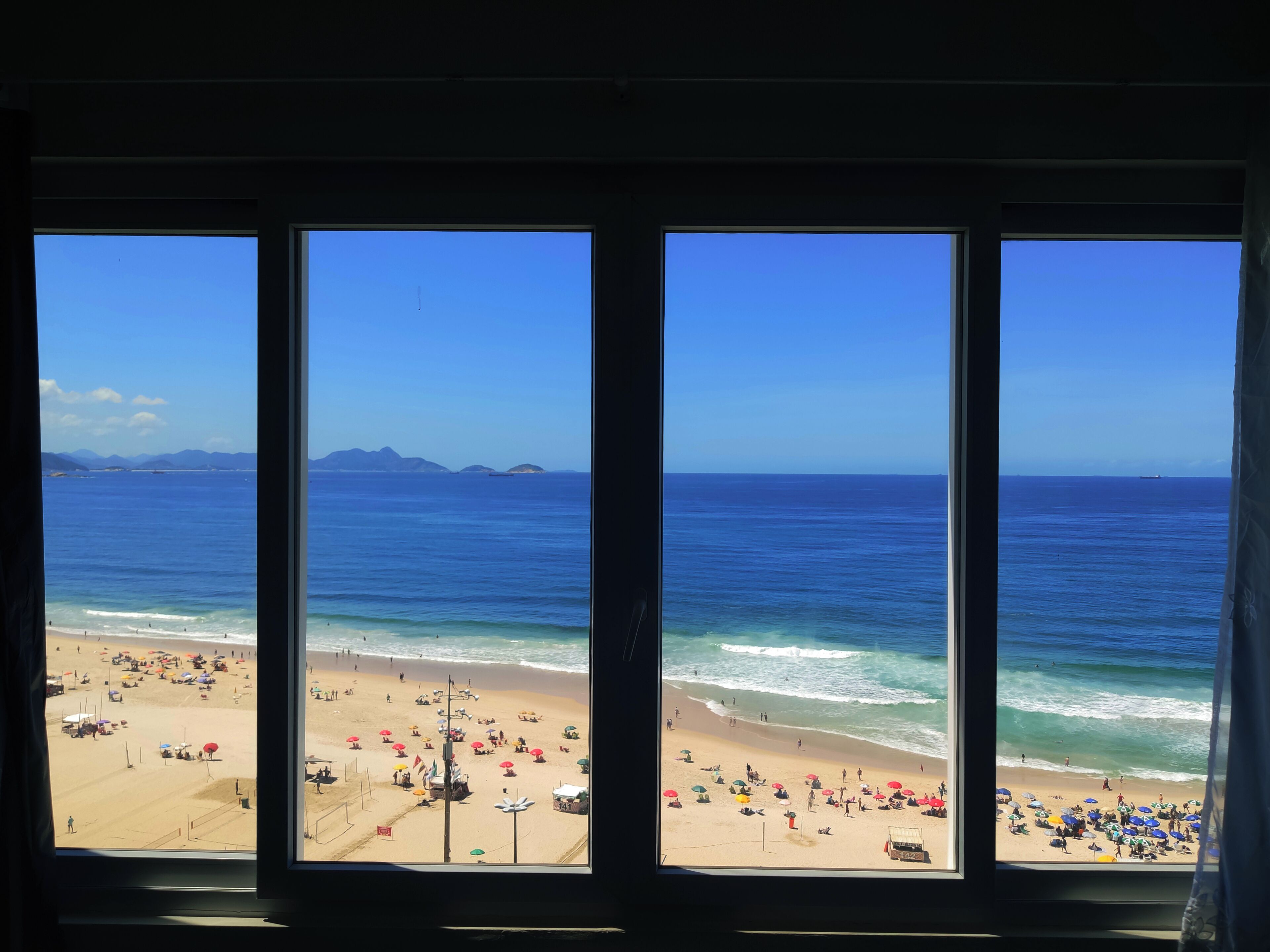 Vista de la playa a través de una ventana, con cielo despejado, mar tranquilo y gente disfrutando en la playa.