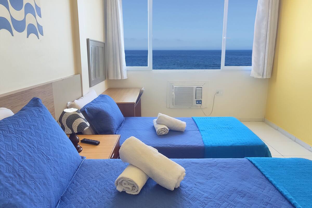 A room with two beds, a TV, a window with a view of the sea, and an air conditioning unit.
