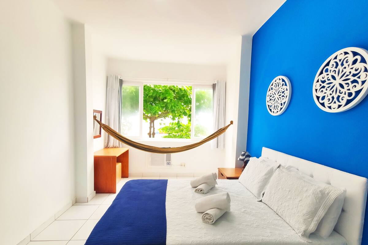 A bedroom with a bed, a hammock, a view of greenery outside, and decorative wall art.