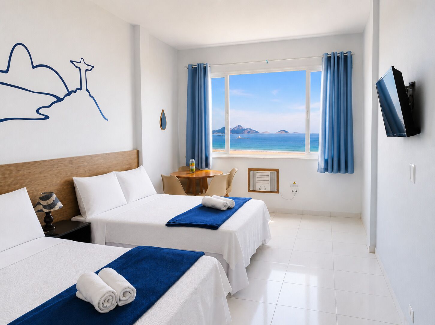 A hotel room with two beds, a desk, a chair, a TV, and a window with a view of the sea.
