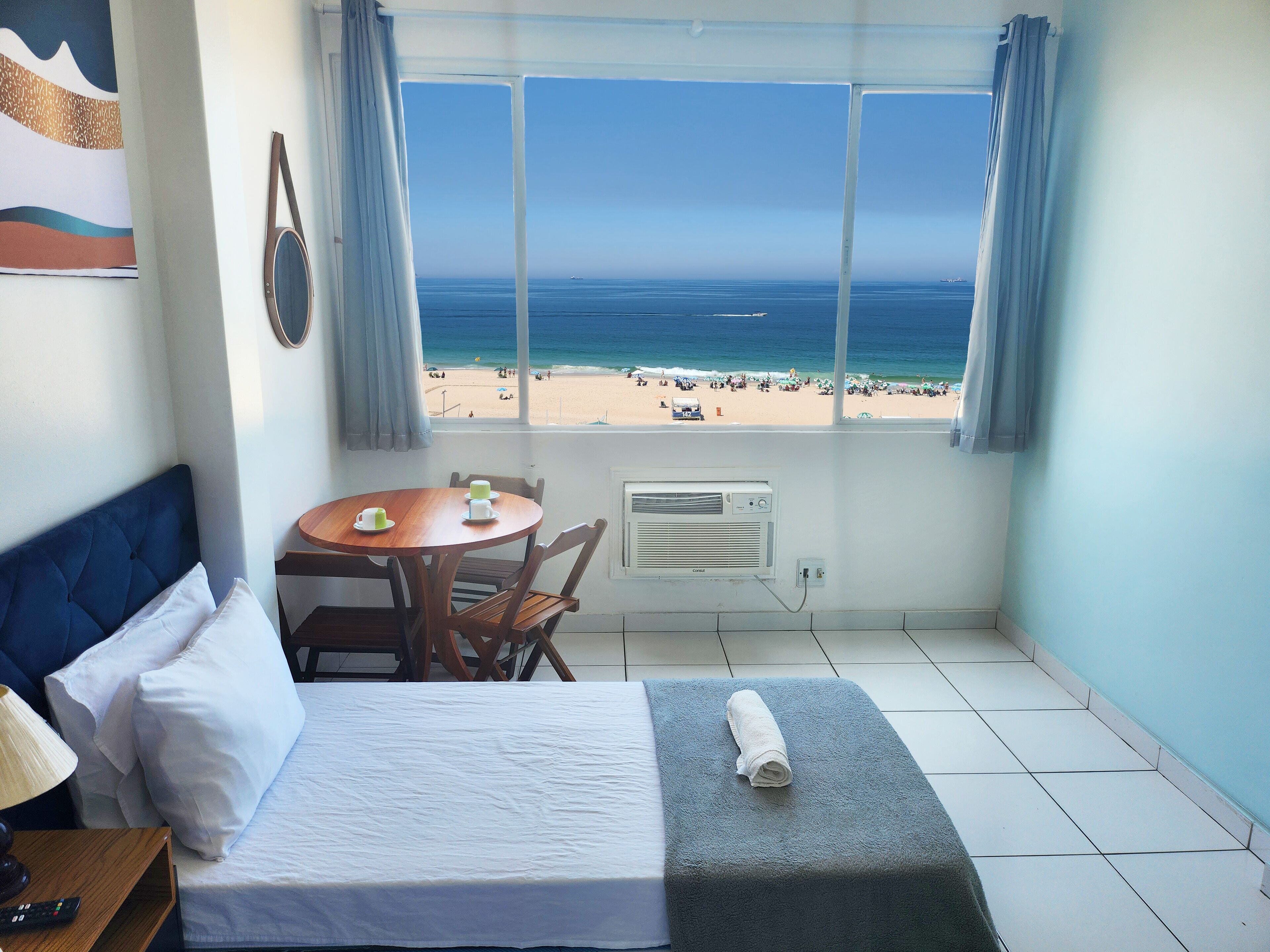 A room with a bed, a table with cups, a view of a beach, and a window with curtains.