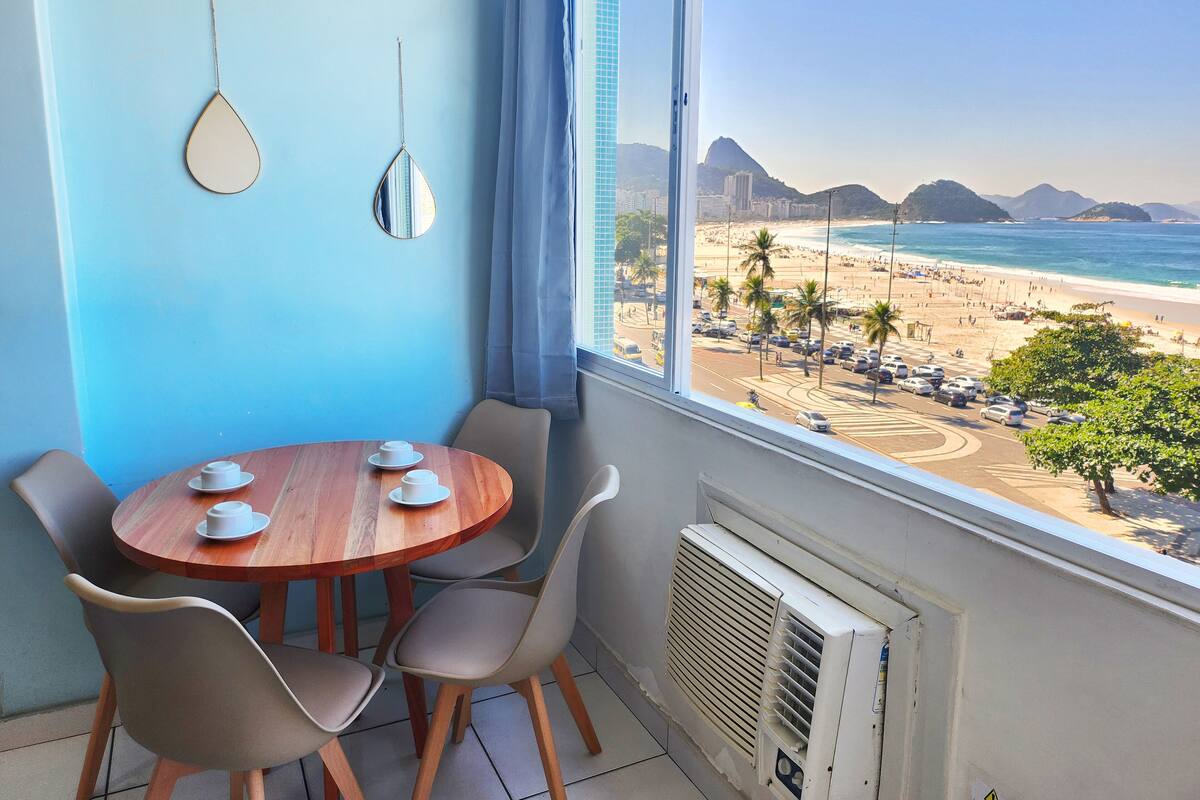A room with a wooden table and chairs, a window with a view of a beach and mountains, and a wall-mounted air conditioner.